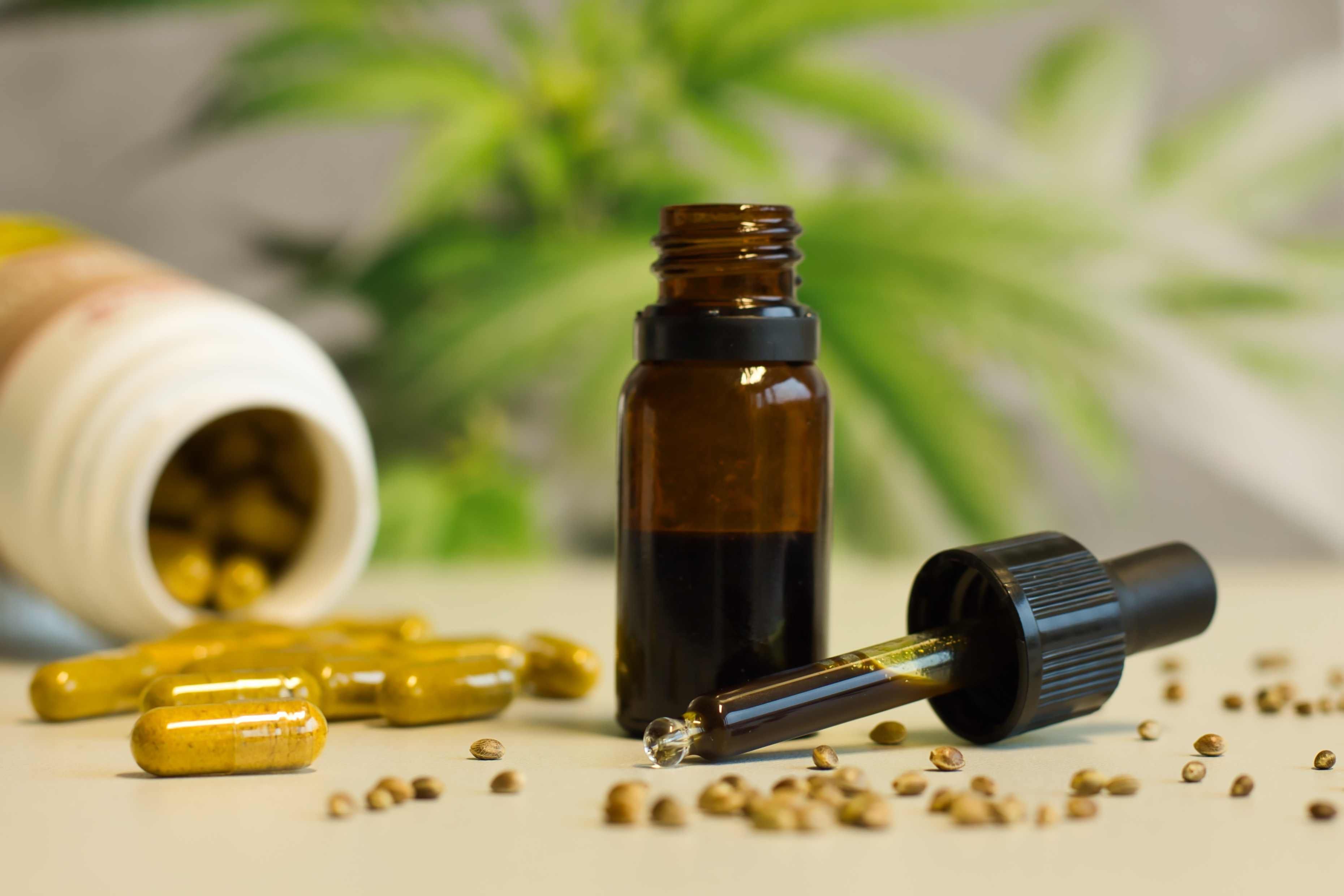 Die Abbildung zeigt eine schwarze 10-ml Glasflasche mit einem CBD-Öl. Die Tropfpipette ist von der Flasche abgeschraubt und liegt gefüllt mit dem dunkel-grünlichen Öl im Vordergrund vor der Flasche. Neben der Flasche liegt ein Plastikbehältnis mit Kapseln, das von oben geöffnet ist. Vor dem Behältnis sind einige ockergelbfarbene Steck-Kapseln platziert, die ein Pulver mit Hanfextrakt enthalten. Weiterhin sind einige kleine Hanfsamen abgebildet, die bräunlich und rund aussehen. Im Hintergrund des Bildes befindet sich ein unscharfes Hanfblatt.